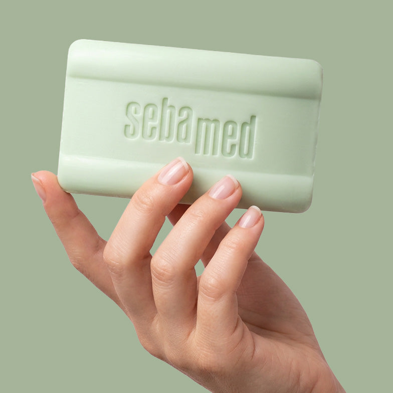 A hand with pale pink manicured nails holds a light sage green Sebamed cleanisng bar against a solid sage green background.