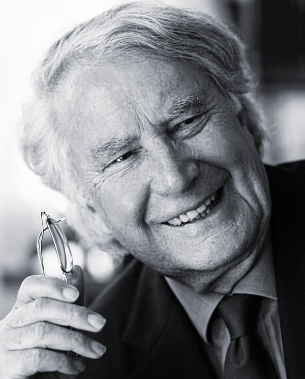 A black and white close-up portrait of Dr Heinz Maurer smiling while holding a pair of eyeglasses, wearing a dark suit jacket, collared shirt, and tie.