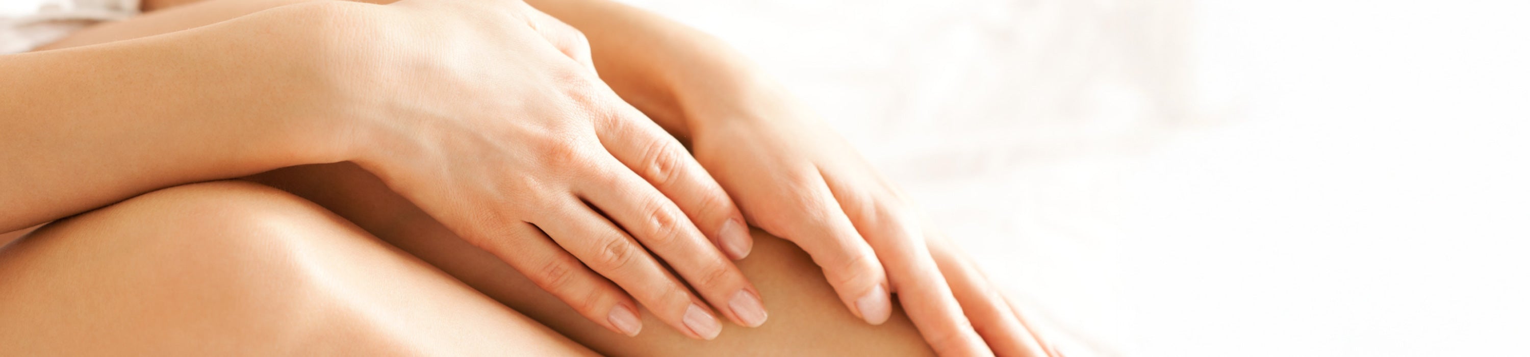 A close-up of a person's hands resting gently on their legs against a bright, softly lit background.
