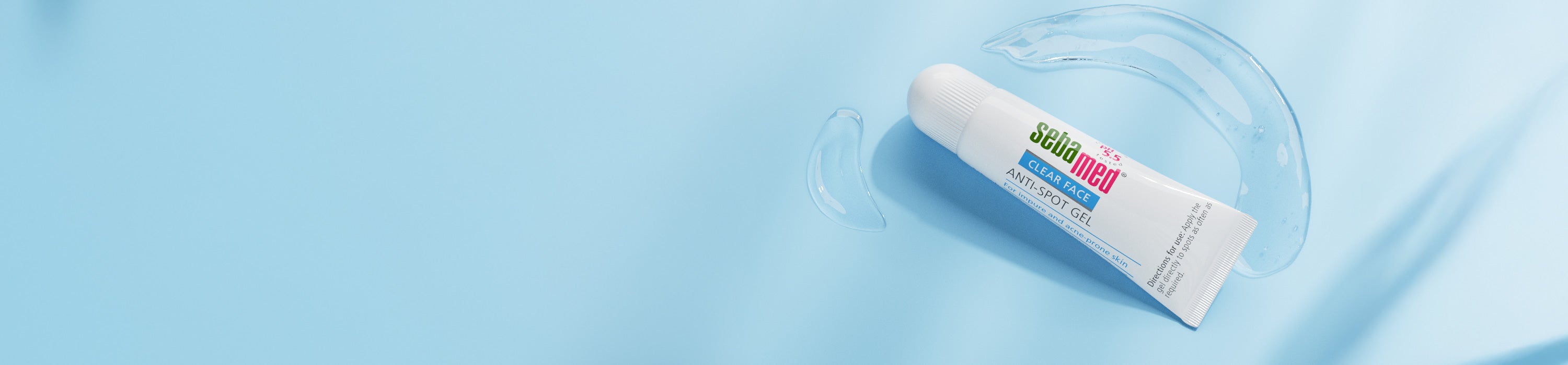 A white squeeze tube of sebamed Anti-Spot Gel, resting on a light blue background with a clear liquid splash of gel around it.