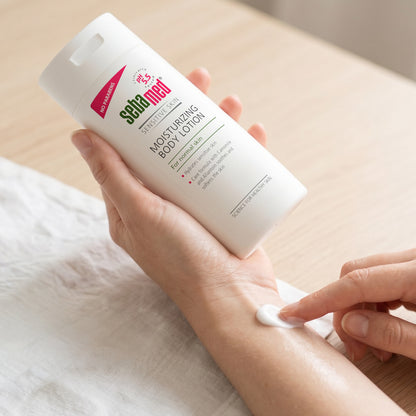 A person holds a white bottle of Sebamed Moisturising Body Lotion over their forearm, with a dollop of the white lotion being spread onto the skin.