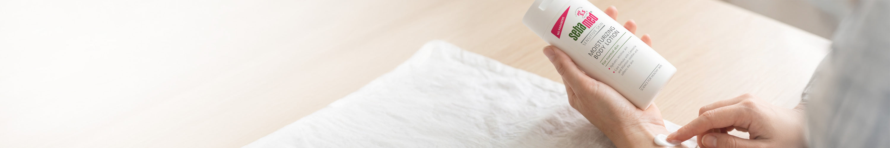 A person holding a white bottle of Sebamed Moisturising Body Lotion over a white towel, with a blurred light background.