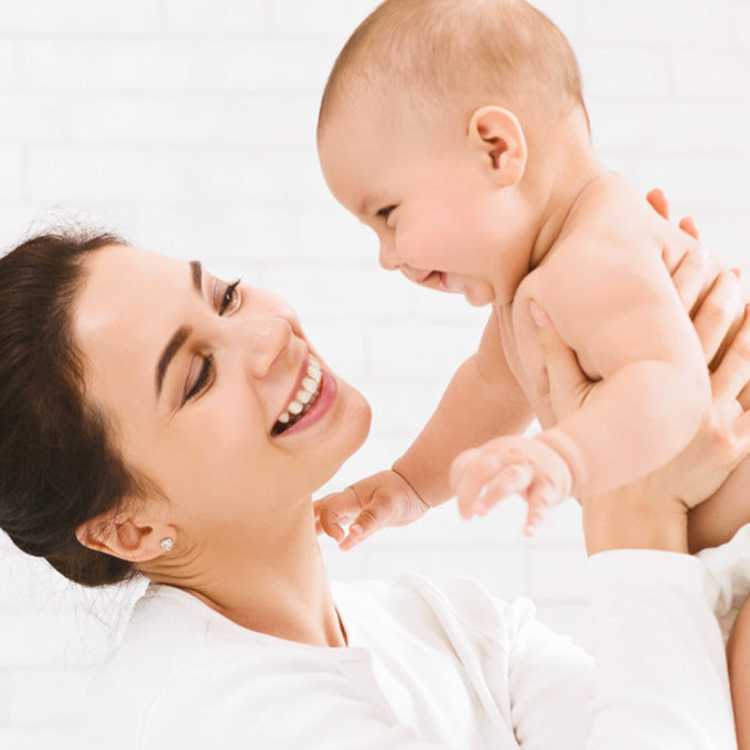 A smiling woman in a white top holds and looks at a baby against a bright white background.