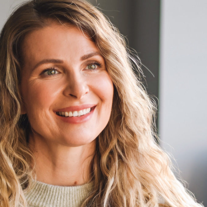 A close-up portrait of a smiling middle-aged woman with long wavy blonde hair, wearing a light beige knit top, set against a soft-focus indoor background.