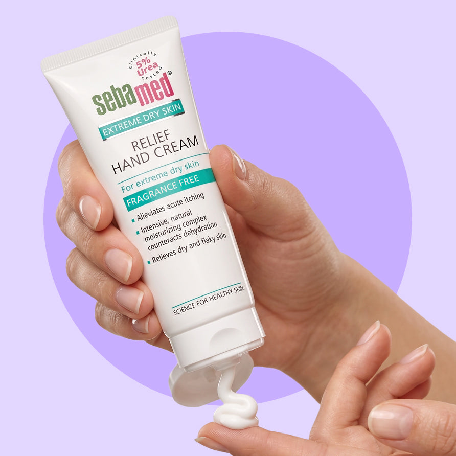 A hand holding a squeeze tube of Sebamed Extreme Dry Skin Relief Hand Cream, with white cream being dispensed onto a surface against a light purple background with a darker purple circular graphic.
