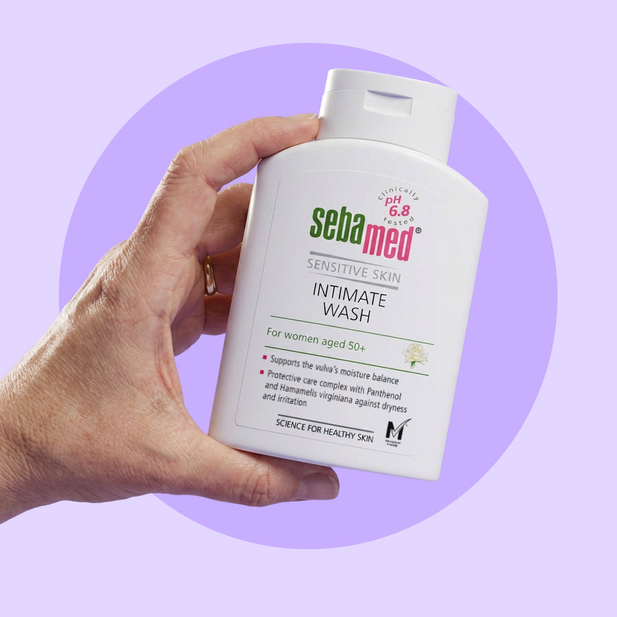 Hand holding a bottle of Sebamed Intimate Wash against a purple background